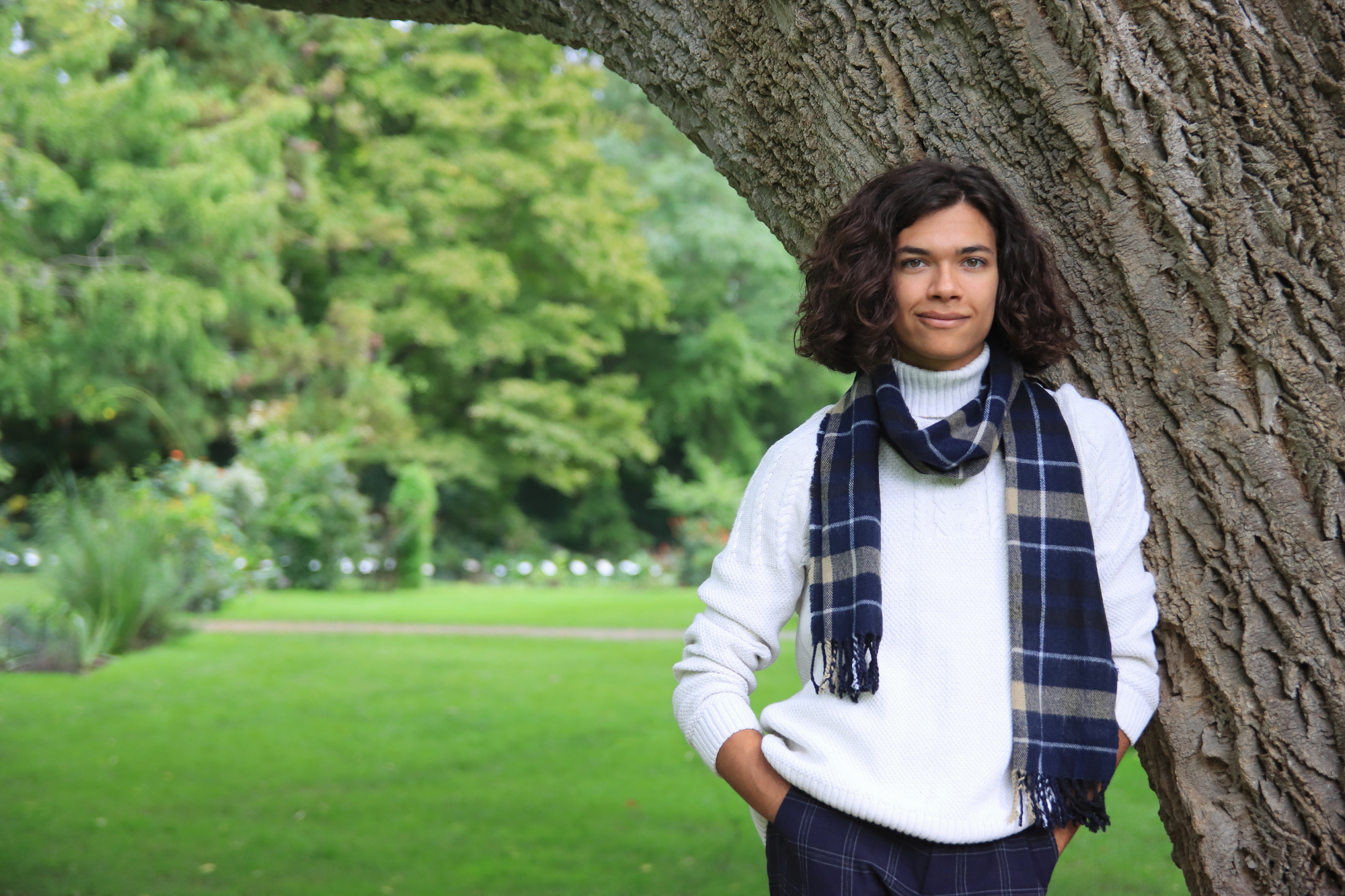 young person leaning against a tree wearing a white sweater and blue scarf