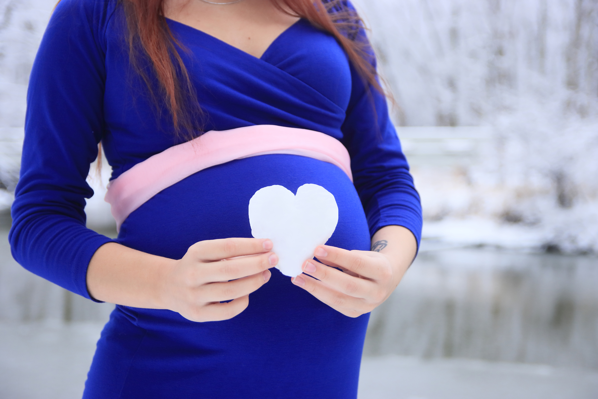 a pregnant woman's belly in a dark blue dress, she has a heart made out of snow resting on her belly