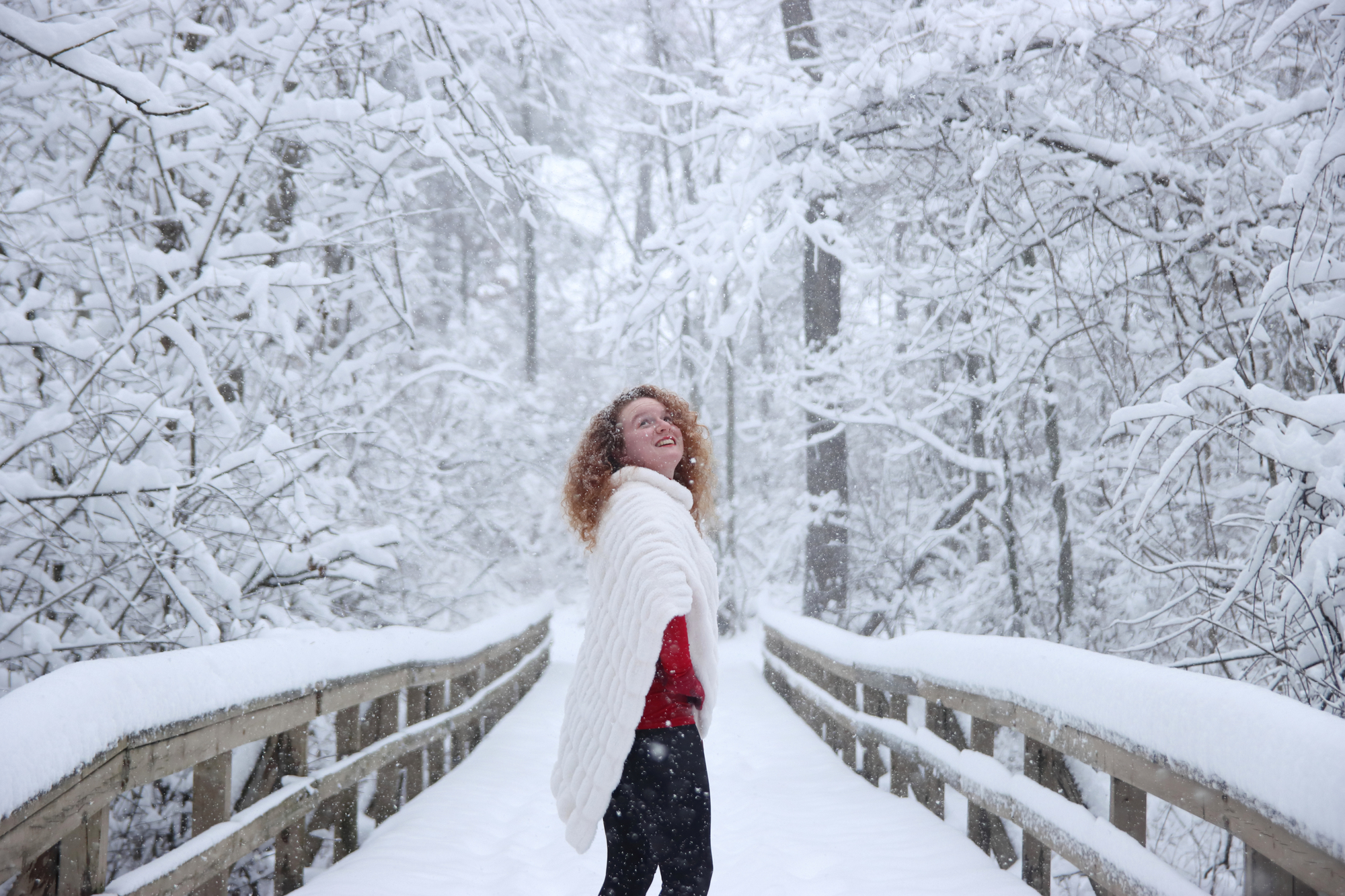 A young woman with bright red hair wearing a bright red outfit poses on a bridge during a snow storm. The contrasting colors are
