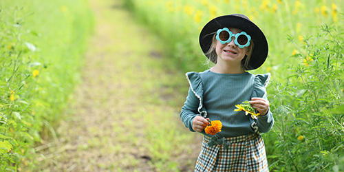 little girl with a black hat and green blue sunglasses smiling in a field of flowers