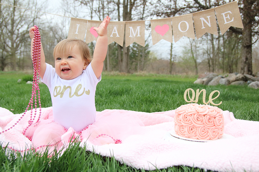 toddler girl sitting on a cream blanket on the grass wearing a pink tutu and a white t-shirt with gold writing that says ONE