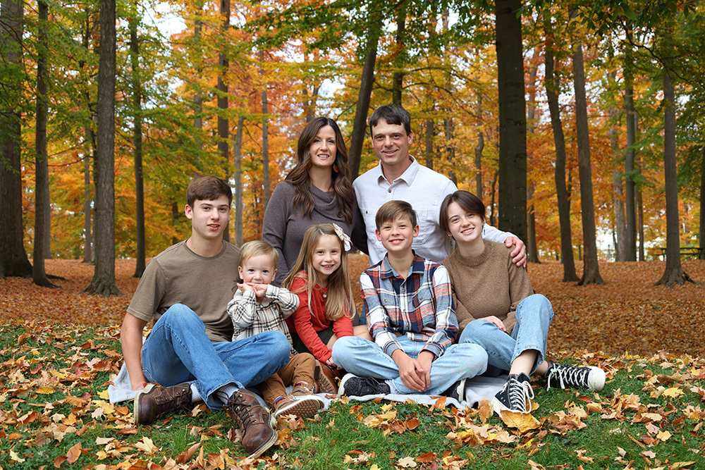 Family of seven posing on a hill with fall trees behind them.