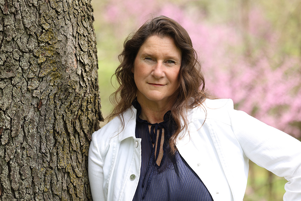 Ariniko O'Meara headshot wearing a blue shirt and white jacket with light pink flowers in the background